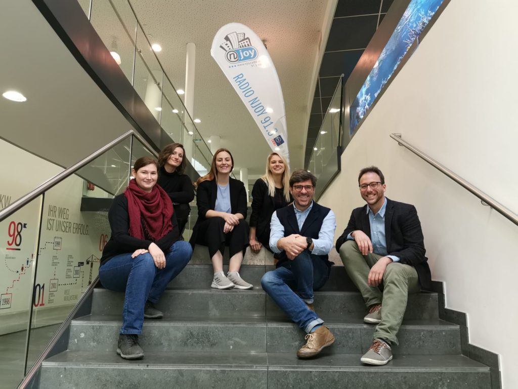 Five young researchers and Andreas Jäger sitting on stairs in front of the Radio NJOY flag. 